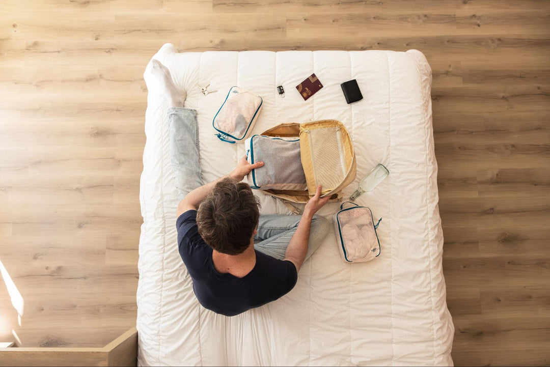 Traveller arranging packing cubes and travel essentials beside an open backpack on a bed.